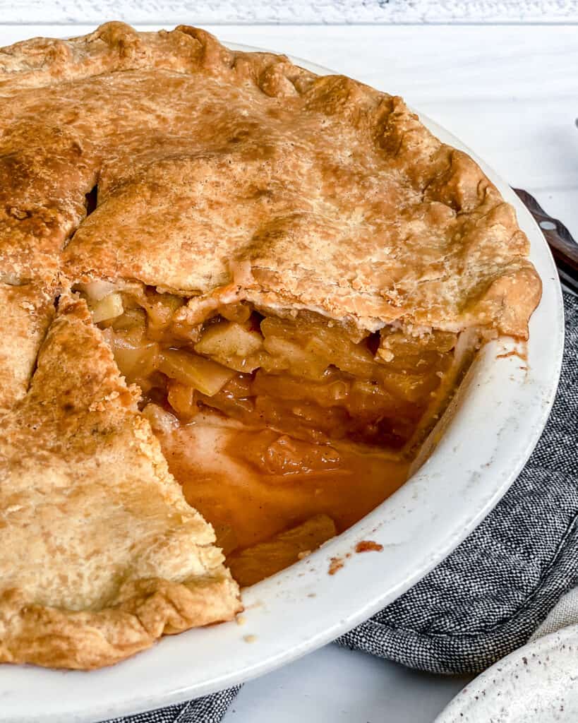 finished apple pie close up in a white pie tray against a white background with a piece missing