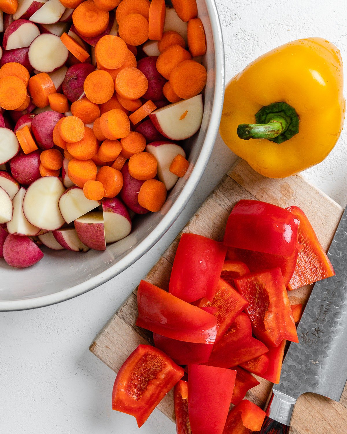 process of cutting peppers on cutting board alongside bowl of potatoes and carrots and yellow pepper