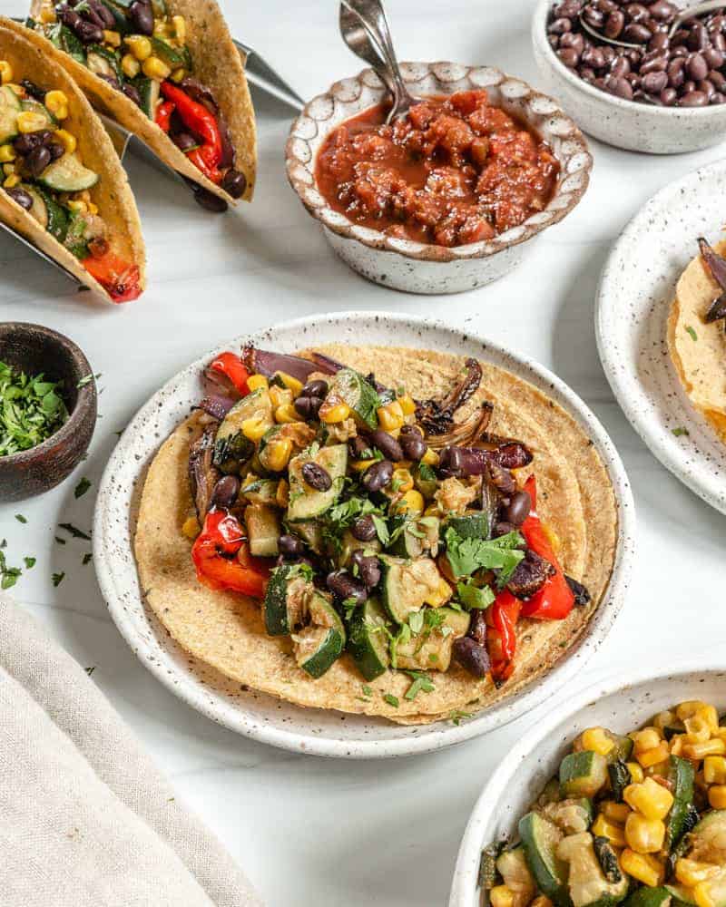 Vegan Roasted Veggie Tacos in a white plate with ingredients in the background against a white surface