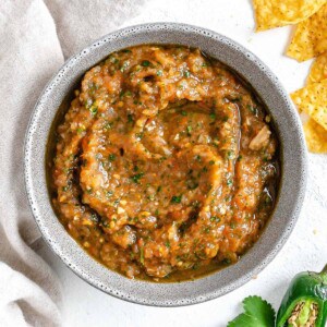 completed Homemade Roasted Red Salsa in a white bowl against a white background with chips an jalapeños in the background