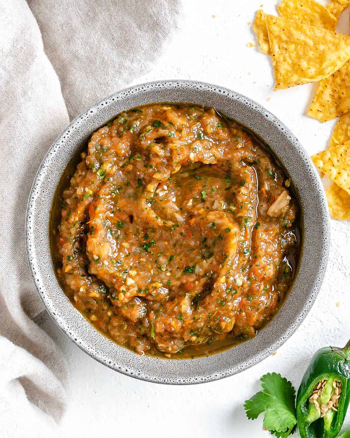 completed Homemade Roasted Red Salsa in a white bowl against a white background with chips an jalapeños in the background 