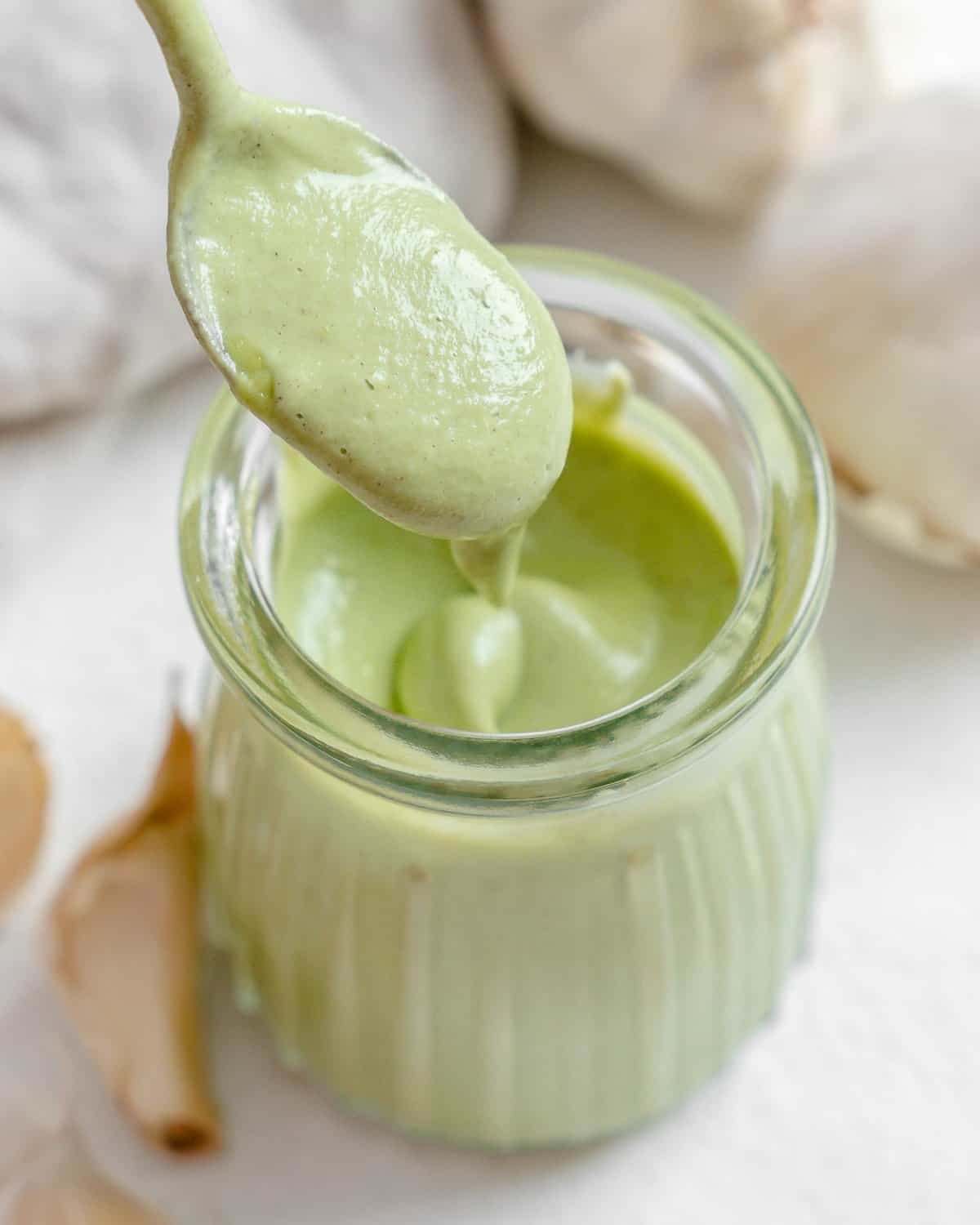 completed Creamy Vegan Garlic Sauce in a glass jar with a spoonful showing consistency with garlic in the background