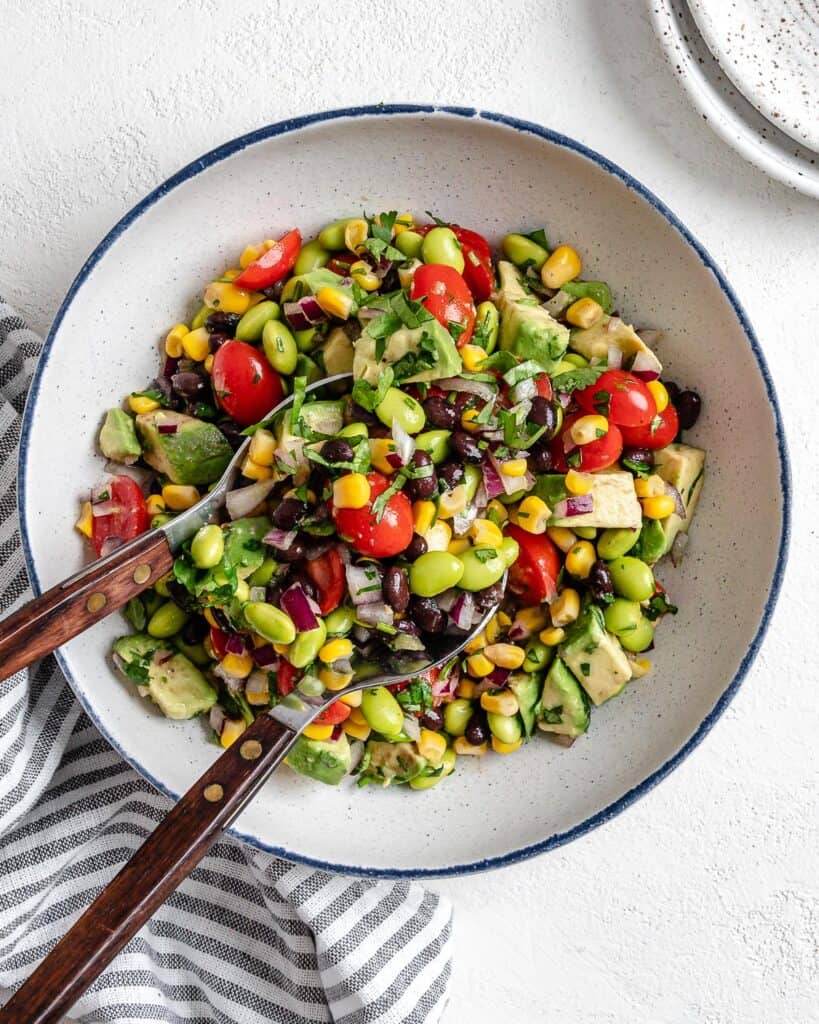 completed white bowl of Easy Edamame Salad with utensils in bowl against a white background