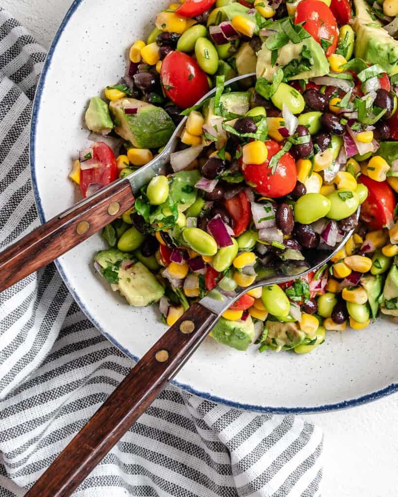 completed white bowl of Easy Edamame Salad with utensils in bowl against a white background