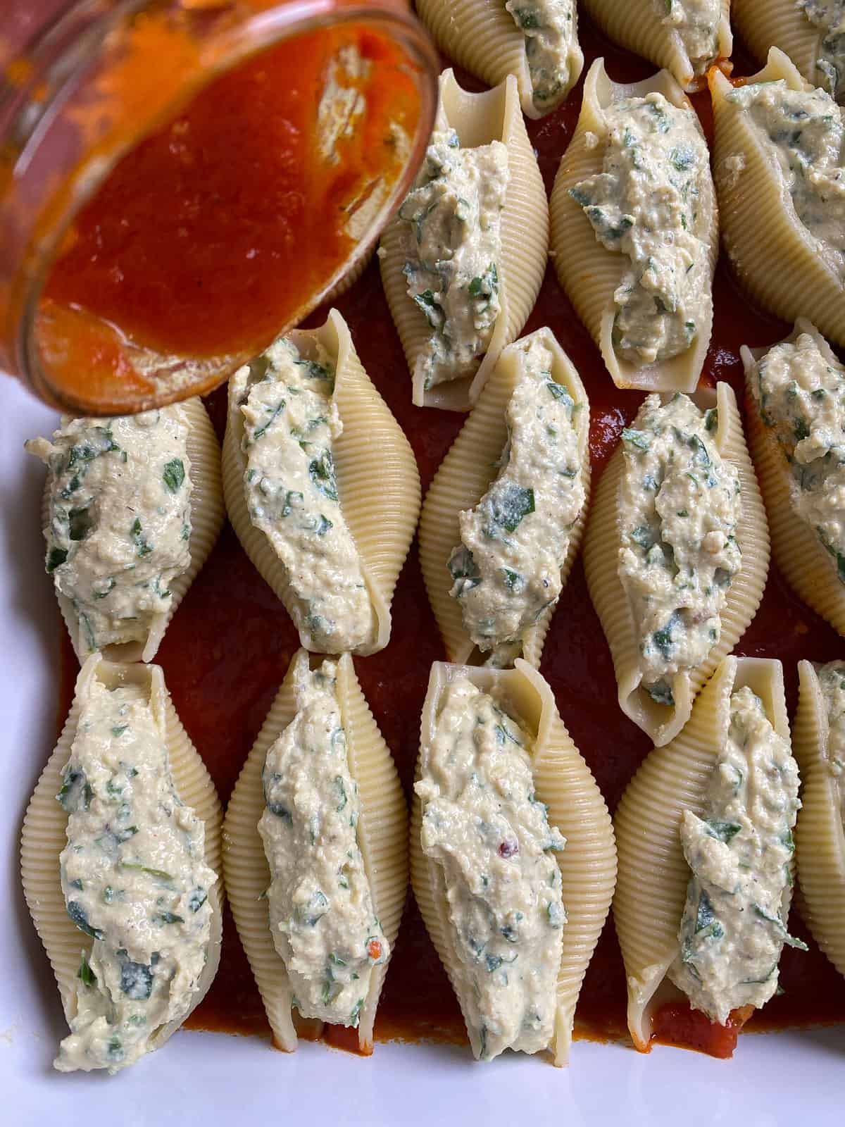 process shot of marinara sauce being drizzled on top of pre baked stuffed shells