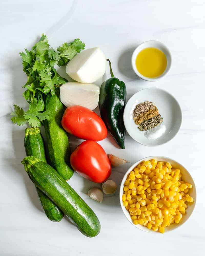 calabasitas ingredients including zucchinis, tomatoes, cilantro, onion, pepper, garlic, corn, and spices in a white marble background