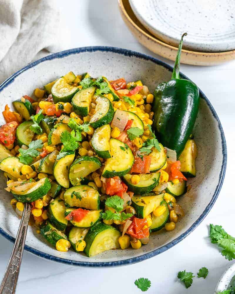 completed calabacitas in a white bowl against a white background with scattered green veggies