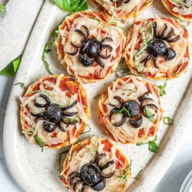 several cooked halloween spider bagel bites on a white platter with ingredients in the background