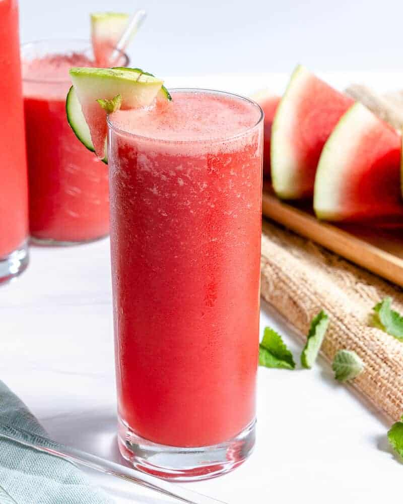 watermelon cooler in tall glass cup with sliced watermelon and more cups of watermelon cooler in a white background