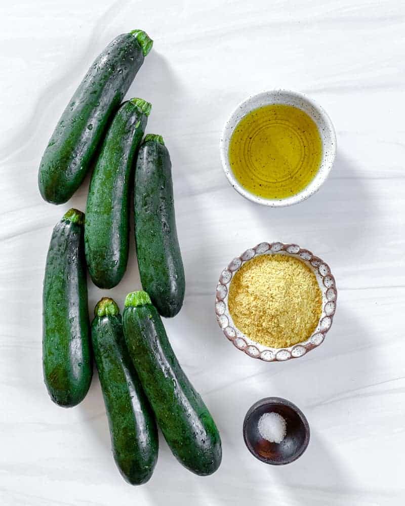 whole zucchini and various ingredients in bowls for vegan cheesy zucchini chips against a white background