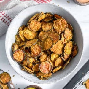 cooked zucchini chip sin a white bowl with zucchini chips in the background