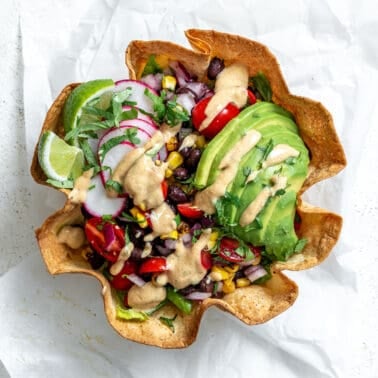 A tortilla bowl holding a taco salad that's topped with a sliced avocado, taco salad dressing, sliced radishes, and lime wedges