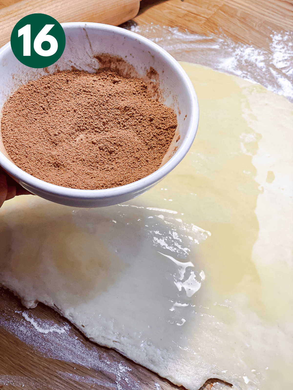 holding a bowl of cinnamon sugar above rolled-out cinnamon roll dough.