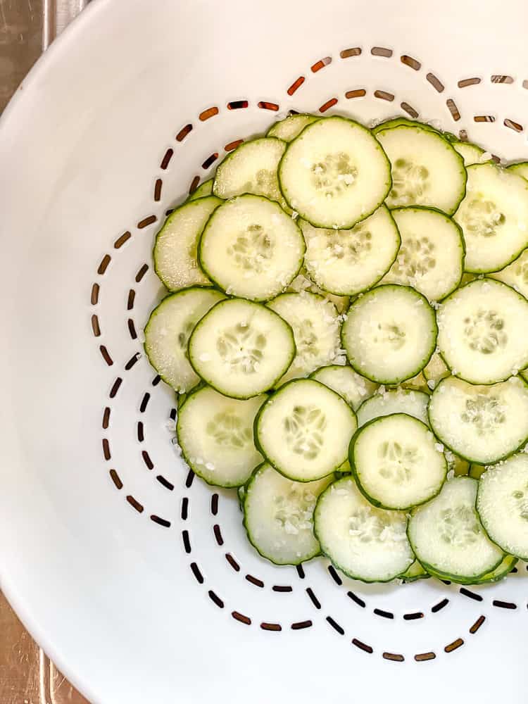 sliced cucumbers topped with salt in a white colander.