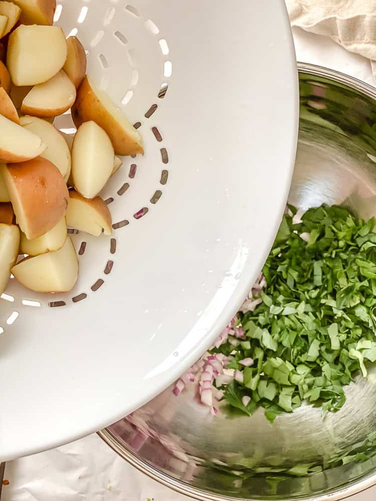 pouring cooked potatoes into a metal bowl with chopped red onion and parsley.