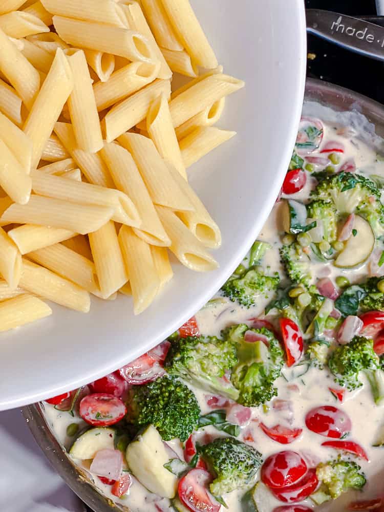 pouring a bowl of cooked pasta into a pot with vegetables and cream sauce.